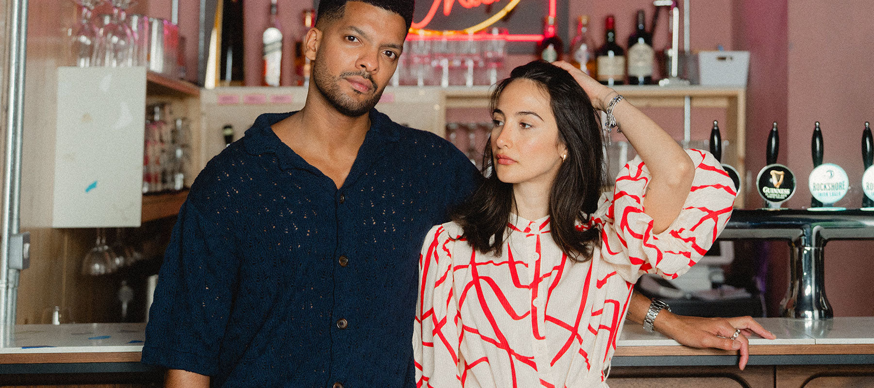 Two people standing in a bar setting with neon signs and bottles in the background. The man is wearing a Knit Shirt and the woman is wearing our Seersucker dress.