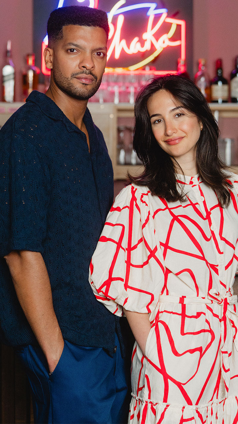Two people standing together in a bar setting with neon signs in the background. Man is in our Knit Shirt, woman is wearing our Seersucker dress.